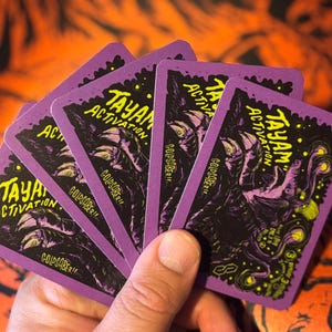 May include: A fan of rectangular playing cards with purple borders, featuring a dark illustration of a creature with glowing yellow eyes and the text TAYAM ACTIVATION and GOLDSABER printed in yellow on a black background with purple accents.