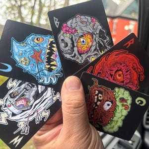 May include: A hand holds a fan of playing cards with grotesque monster illustrations. Each card features a black background with a different colorful, detailed design of a monster face. The cards are in various suits and values.