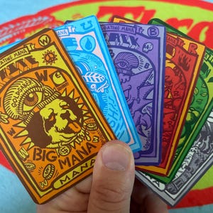 May include: A hand holds a fan of colorful cards with unique illustrations and text. The cards feature various designs, including a dog, an eye, and other symbols, with the words "Floating Mana" and "Big Mana" visible. The cards are in shades of orange, blue, purple, red, green, and gray.