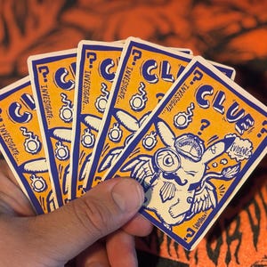 May include: A set of five rectangular playing cards with a yellow background and blue borders. The cards feature a cartoon character wearing a detective hat and mustache, holding a magnifying glass, with the text CLUE and INVESTIGATE.