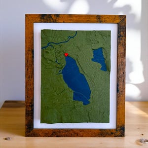 May include: A framed artwork featuring a topographical map with a blue lake and green land. A red heart marks a location on the map. The artwork is set in a wooden frame with a white mat, displayed on a wooden surface.
