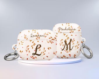 Puede incluir: Dos estuches blancos para Airpods con patrones florales y nombres personalizados. El estuche de la izquierda dice "Lauren" con una "L" marrón y el estuche de la derecha dice "Madeline" con una "M" marrón.