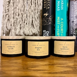 May include: Three black candle jars with wooden lids. The labels are brown with white text. The candles are labeled "Scribes & Scrolls", "Dragons Den", and "Faerie Apothecary".