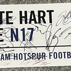 Puede incluir: Un letrero rectangular de metal blanco con el texto negro WHITE HART LANE N17 y TOTTENHAM HOTSPUR FOOTBALL CLUB. El letrero incluye el logotipo azul del gallo de Tottenham Hotspur y está cubierto con numerosas firmas en tinta negra de personas.