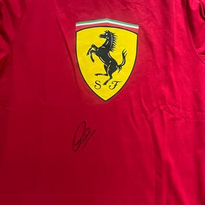 Può includere: T-shirt rosso con il logo Ferrari, un cavallo nero rampante su uno scudo giallo con dettagli verdi e bianchi. La maglietta ha una striscia bianca sulla spalla e una firma nera.