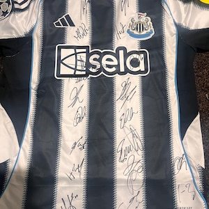 May include: A Newcastle United soccer jersey with black and white stripes, featuring the Adidas logo and the "sela" sponsor. The jersey is signed by multiple people and has a crest on the chest.