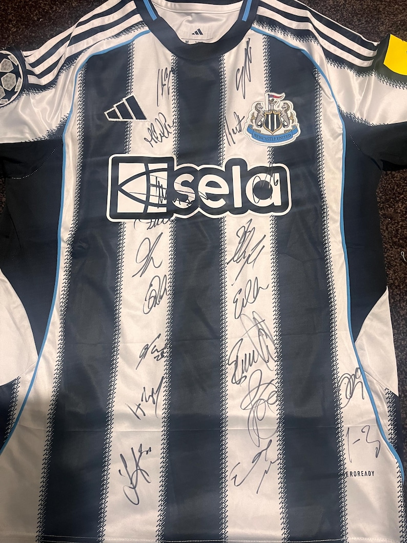 May include: A Newcastle United soccer jersey with black and white stripes, featuring the Adidas logo and the "sela" sponsor. The jersey is signed by multiple people and has a crest on the chest.
