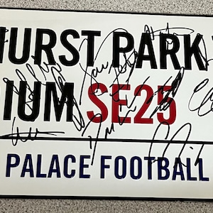 Può includere: Una targa rettangolare in metallo bianco con il testo nero SELHURST PARK STADIUM, il testo rosso SE25 e il testo blu CRYSTAL PALACE FOOTBALL CLUB. La targa include il logo dell'aquila del Crystal Palace F.C. e numerose firme a inchiostro nero.