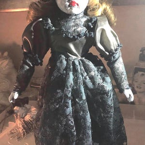 May include: A porcelain doll with blonde hair, wearing a black lace dress and a white hat with a pink ribbon. The doll has a painted face with dark eyes and red lips.