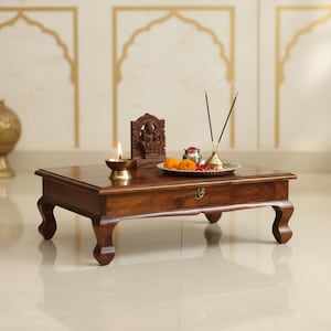 Puede incluir: Una mesa de oración rectangular de madera con cuatro patas curvas y un cajón frontal con tirador de metal. Encima hay una pequeña lámpara de aceite de latón encendida, una figura de deidad tallada en madera y una bandeja plateada con incienso.