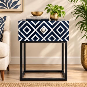 May include: A square bedside table featuring a navy blue and white geometric diamond pattern on the drawer front, a white flower-shaped knob, and a black metal frame. The top surface holds a gold bowl, a plant, and two books titled The Kinfolk Home and Louis Vuitton.