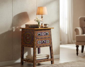 May include: A wooden bedside table with two drawers, featuring a purple finish and intricate floral patterns in pink, green, and white. The table has four legs and a lower shelf holding a folded white cloth. A lamp, books, and a cup sit on top.