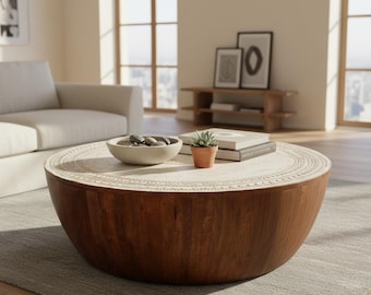 May include: A round, drum-shaped coffee table crafted from dark brown wood with a light-colored, circular top featuring an intricate carved border pattern. The table surface holds a shallow, light-toned bowl filled with stones and a small succulent.