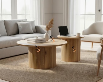 Puede incluir: Dos mesas redondas de madera clara con bases estriadas verticales. La mesa más grande tiene una superficie plana con una pila de libros y un cuenco pequeño, mientras que la mesa más pequeña sostiene un portátil abierto y una taza.