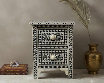May include: A rectangular bedside table featuring two drawers with intricate black and white floral inlay patterns. The piece has a flat top, four short legs, and two round, cream-colored knobs. The surface is decorated with repeating geometric motifs.