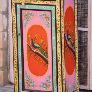 May include: A small, hand-painted wooden cabinet with a single door and drawer. The cabinet is decorated with colorful floral patterns and peacocks. The door is painted a bright pink with a red oval in the center featuring a peacock. The cabinet is painted in shades of pink, yellow, blue, green, and brown.