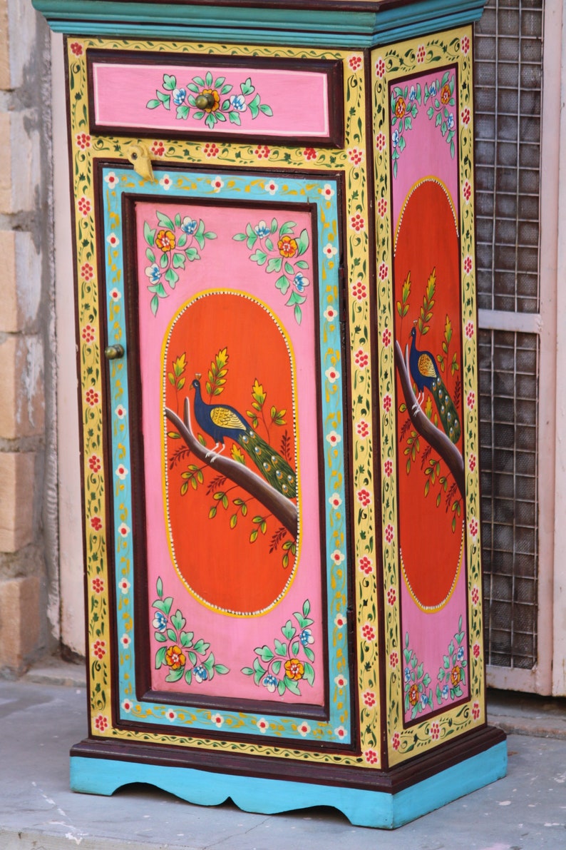 May include: A small, hand-painted wooden cabinet with a single door and drawer. The cabinet is decorated with colorful floral patterns and peacocks. The door is painted a bright pink with a red oval in the center featuring a peacock. The cabinet is painted in shades of pink, yellow, blue, green, and brown.