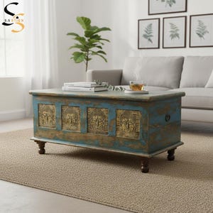 May include: A rectangular wooden coffee table with a distressed blue finish and four carved gold-toned panels featuring Buddha faces. The table stands on four dark brown turned feet and includes metal side handles for lifting the lid.
