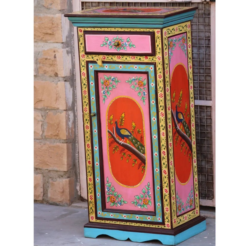 May include: A hand-painted wooden cabinet with a single drawer and a door. The cabinet is decorated with peacocks, flowers, and geometric patterns in bright colors, including pink, orange, yellow, blue, and green.