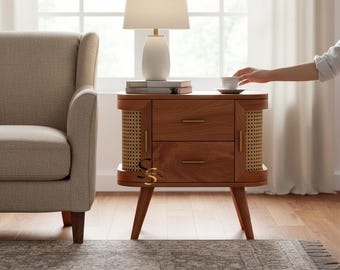 May include: A wooden bedside table with two drawers, gold-toned horizontal handles, and rounded sides featuring woven cane panels. The table stands on four tapered legs and supports a white table lamp with a cylindrical base and fabric shade.