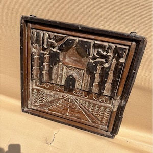 May include: A square wooden wall plaque featuring a hand-carved relief of the Taj Mahal. The dark brown wood displays intricate architectural details, including the central dome, arched entrance, and surrounding minarets, with a path leading forward.