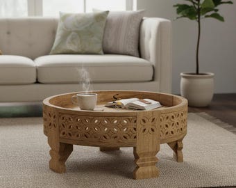 May include: A round, light brown wooden coffee table featuring intricate, carved floral cutouts along its side panels and four sturdy, carved legs. The table surface holds an open book and a steaming white ceramic mug of hot beverage.