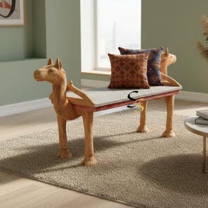 May include: A wooden bench featuring carved camel heads at each end, with a flat seat covered in a light-colored patterned fabric. Two square throw pillows are placed on the seat, one in a rust-orange geometric print and one in a dark blue pattern.