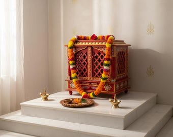 May include: A red wooden shrine with intricate lattice doors and gold accents, adorned with a vibrant marigold garland. Two brass oil lamps flank a circular wooden tray containing small offerings, all placed upon a tiered white marble platform.