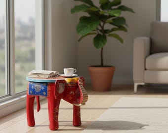 May include: A small, red, elephant-shaped side table with a round, light blue top. The table features intricate, colorful painted patterns in blue, yellow, and white on its sides and legs. An open book and a white coffee cup sit on the table surface.