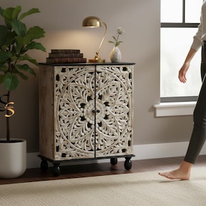 Puede incluir: Un gabinete rectangular de madera con dos puertas que presentan intrincados patrones florales de mandala tallados a mano en un acabado blanco desgastado. El mueble tiene una base y una parte superior negras sólidas, apoyadas sobre cuatro patas negras redondeadas.