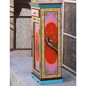 May include: A tall, narrow cabinet with a single door and a small drawer. The cabinet is painted in bright colors, including pink, orange, yellow, green, and blue. A peacock is painted on the door, and the cabinet is decorated with floral designs.