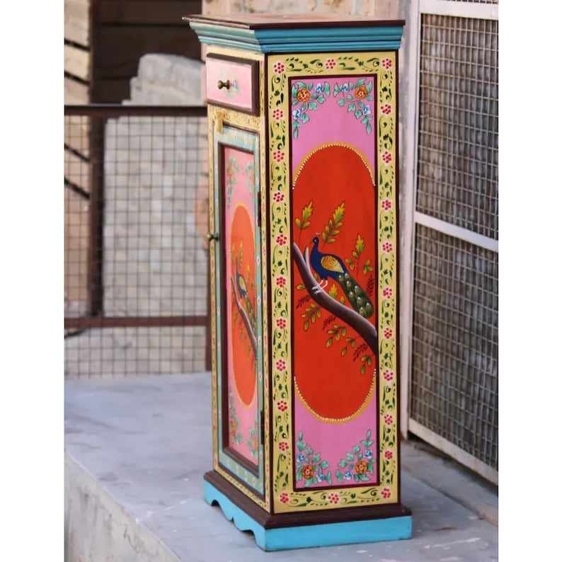 May include: A tall, narrow cabinet with a single door and a small drawer. The cabinet is painted in bright colors, including pink, orange, yellow, green, and blue. A peacock is painted on the door, and the cabinet is decorated with floral designs.