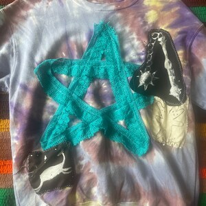 May include: A tie-dye sweatshirt with various patches sewn on. The patches include a large teal star, a black and white skeleton, a white cat, and a beige leaf.