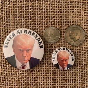 May include: Two round button pins with a white background and black text that reads "Never Surrender". The buttons feature a photo of Donald Trump. The larger button is approximately 2 inches in diameter, and the smaller button is approximately 1 inch in diameter.