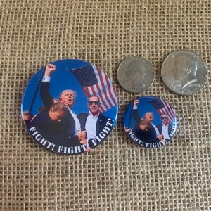 May include: Two round buttons with an image of three people, including Donald Trump, with the text "Fight! Fight! Fight!" printed on the bottom. The buttons are on a brown burlap background.