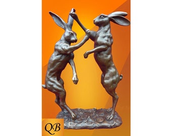 May include: Bronze sculpture of two hares boxing, standing on a textured base. The sculpture is on a bright orange background.