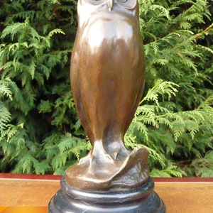 May include: A bronze owl sculpture with a smooth, polished finish. The owl has large, round eyes and is perched on a black, circular base. The sculpture is set against a backdrop of green foliage.
