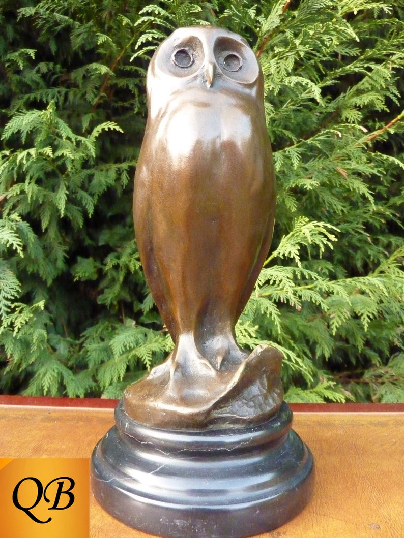 May include: A bronze owl sculpture with a smooth, polished finish. The owl has large, round eyes and is perched on a black, circular base. The sculpture is set against a backdrop of green foliage.
