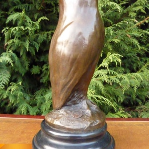 May include: A bronze owl sculpture with a dark patina. The owl is perched on a black, round base. The sculpture has a smooth, polished finish and is set against a backdrop of green foliage.