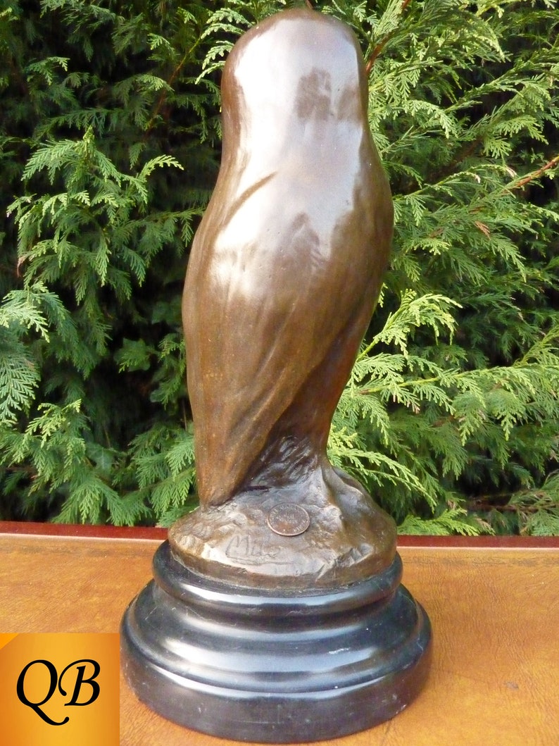 May include: A bronze owl sculpture with a dark patina. The owl is perched on a black, round base. The sculpture has a smooth, polished finish and is set against a backdrop of green foliage.