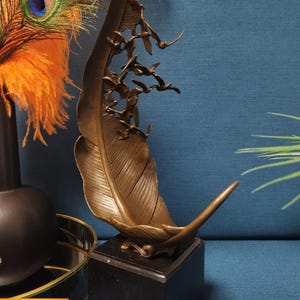 May include: Bronze feather sculpture with a flock of birds in flight, mounted on a black marble base. The sculpture has intricate details and a warm, metallic finish. Decorative art piece for home decor.