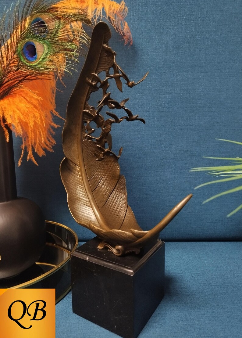 May include: Bronze feather sculpture with a flock of birds in flight, mounted on a black marble base. The sculpture has intricate details and a warm, metallic finish. Decorative art piece for home decor.