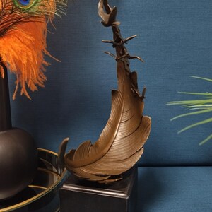 May include: Bronze feather sculpture with a branch and birds, mounted on a black square base. The feather has detailed texture and is set against a blue background. Decorative art.