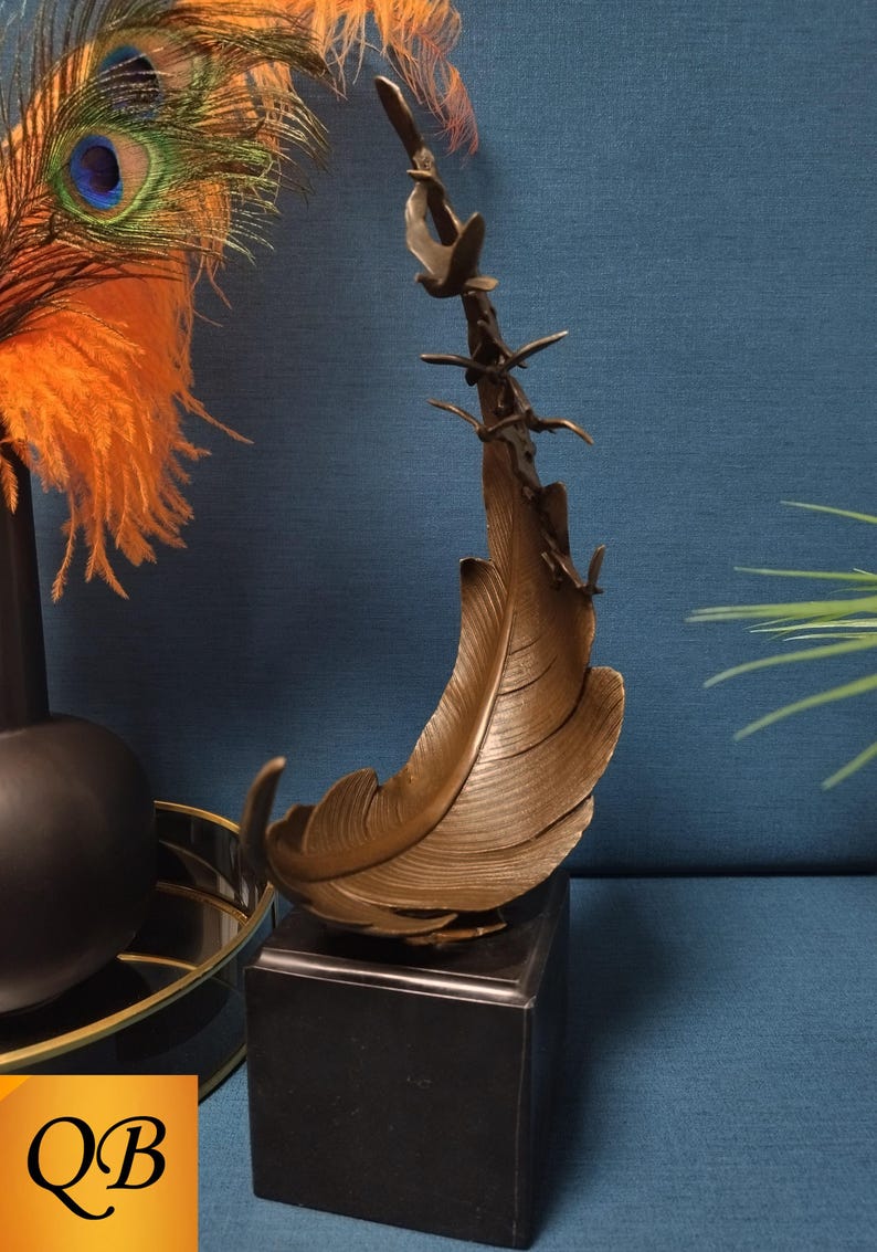 May include: Bronze feather sculpture with a branch and birds, mounted on a black square base. The feather has detailed texture and is set against a blue background. Decorative art.