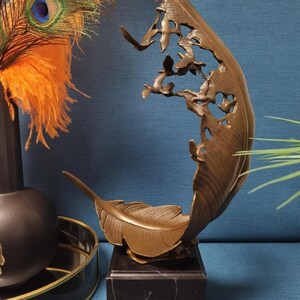 May include: Bronze feather sculpture with a cutout design of birds, mounted on a black marble base. The sculpture has a curved shape and detailed feather texture. Decorative peacock feathers and a black vase are in the background.