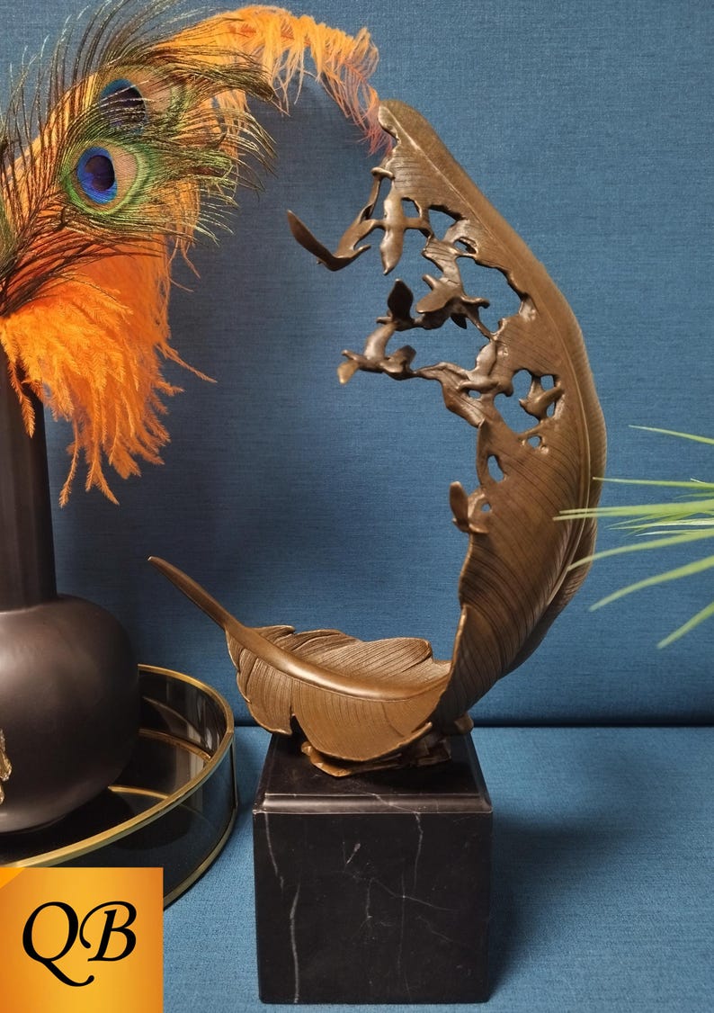 May include: Bronze feather sculpture with a cutout design of birds, mounted on a black marble base. The sculpture has a curved shape and detailed feather texture. Decorative peacock feathers and a black vase are in the background.