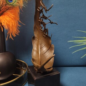 May include: Bronze feather sculpture with flying birds, mounted on a black square base. The feather has detailed veining and a textured surface. Decorative art piece for home decor.