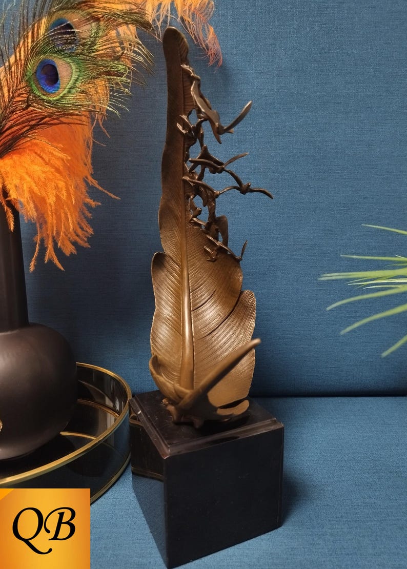 May include: Bronze feather sculpture with flying birds, mounted on a black square base. The feather has detailed veining and a textured surface. Decorative art piece for home decor.