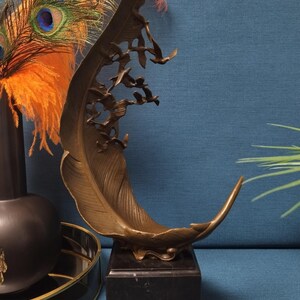 May include: Bronze feather sculpture with flying birds, mounted on a black marble base. The sculpture has a curved shape and intricate details. Decorative accents include peacock feathers and a black vase.