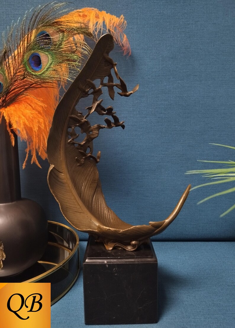 May include: Bronze feather sculpture with flying birds, mounted on a black marble base. The sculpture has a curved shape and intricate details. Decorative accents include peacock feathers and a black vase.